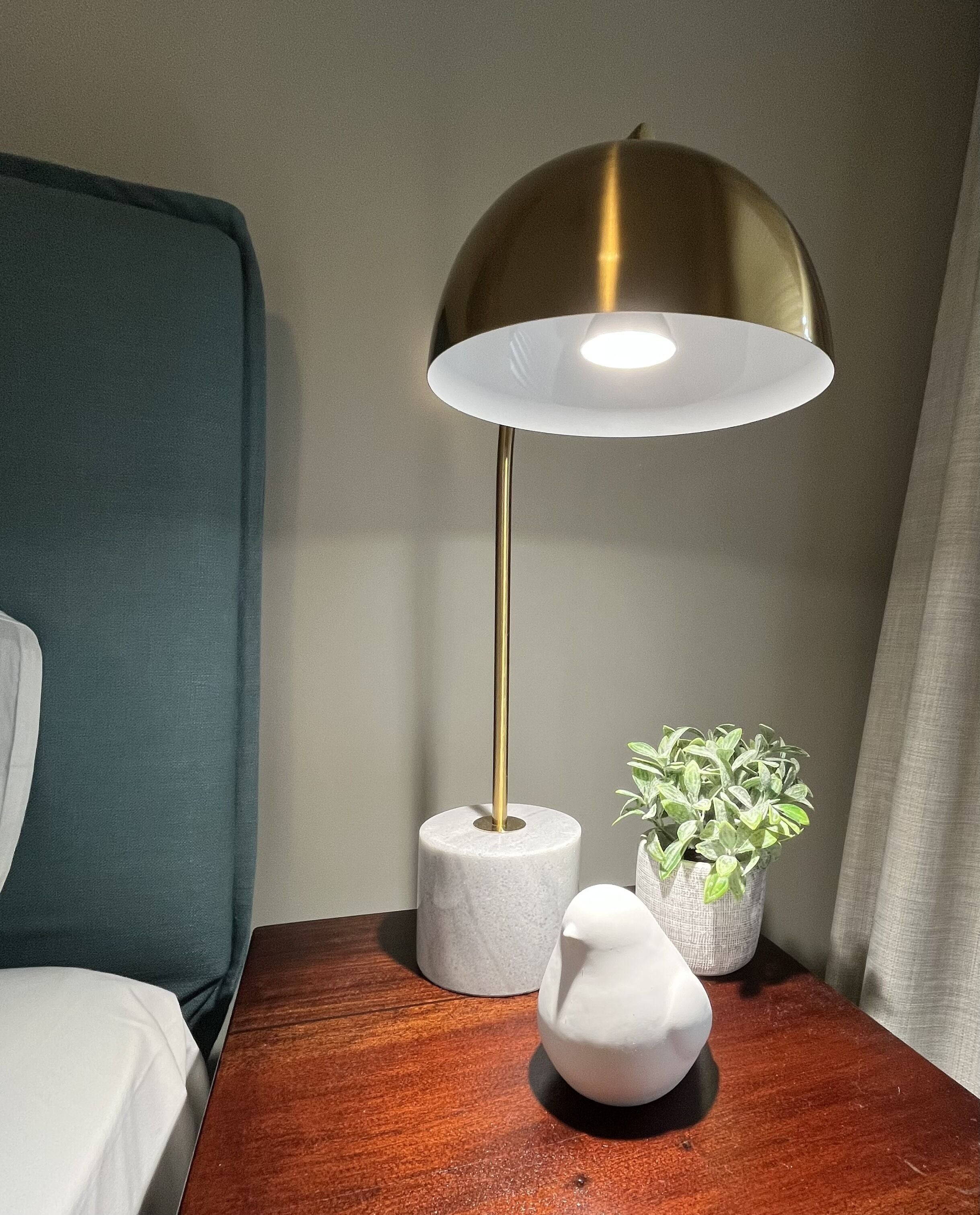 A modern room with a wooden nightstand, a green headboard, a white pillow, a white lamp with a gold base, a white vase, and a potted plant.