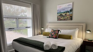 Un lit bien fait, avec du linge de lit blanc et vert, un coussin à motifs floraux et des serviettes enroulées.