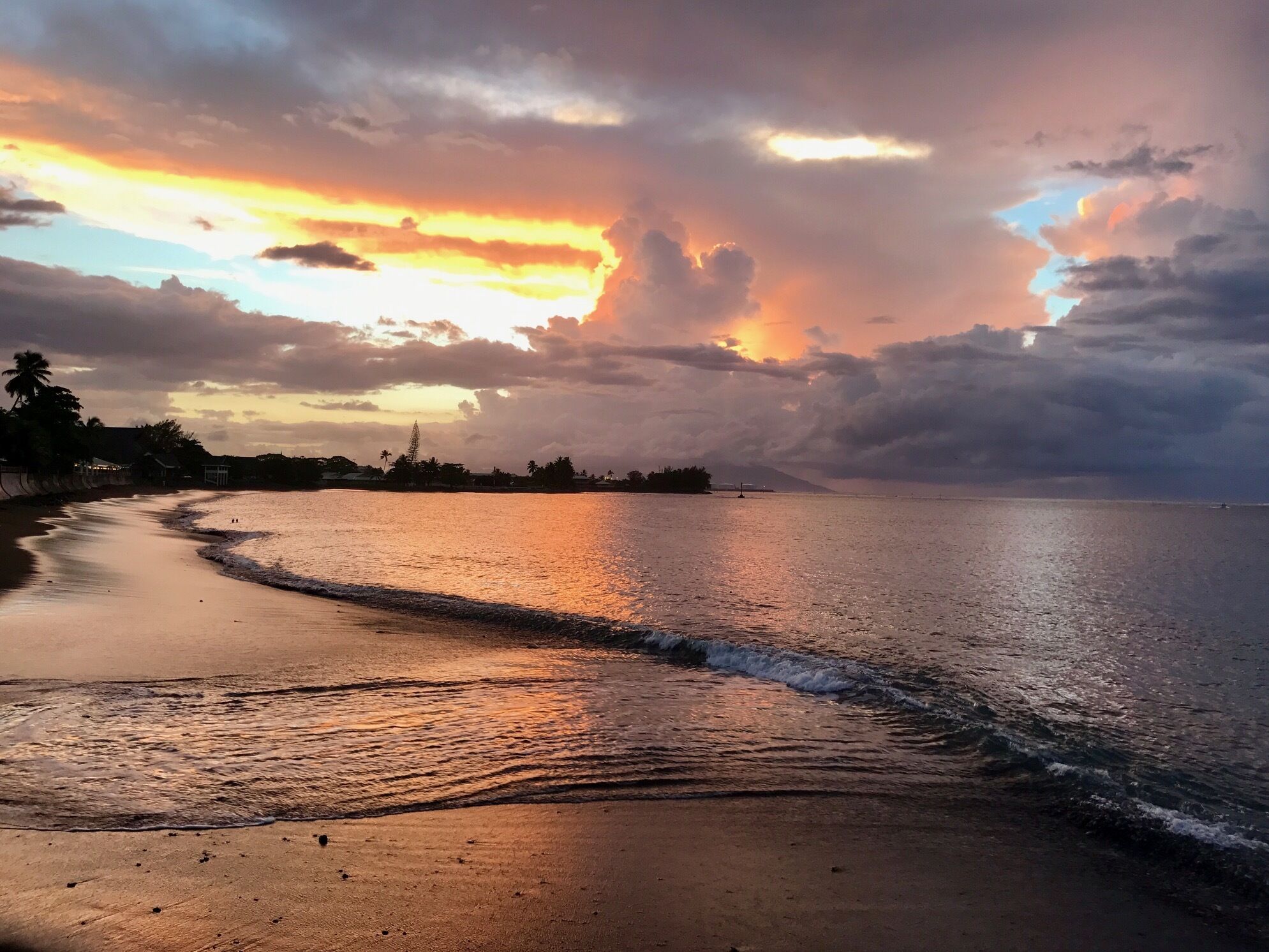 Une plage au coucher du soleil, avec une eau calme et une côte lointaine parsemée de bâtiments et de palmiers.