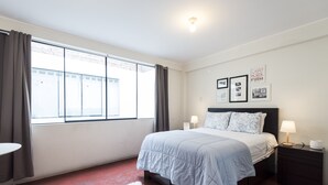 A bedroom with a large bed, a nightstand, a bedside table, a window with curtains, and a wall with framed pictures.