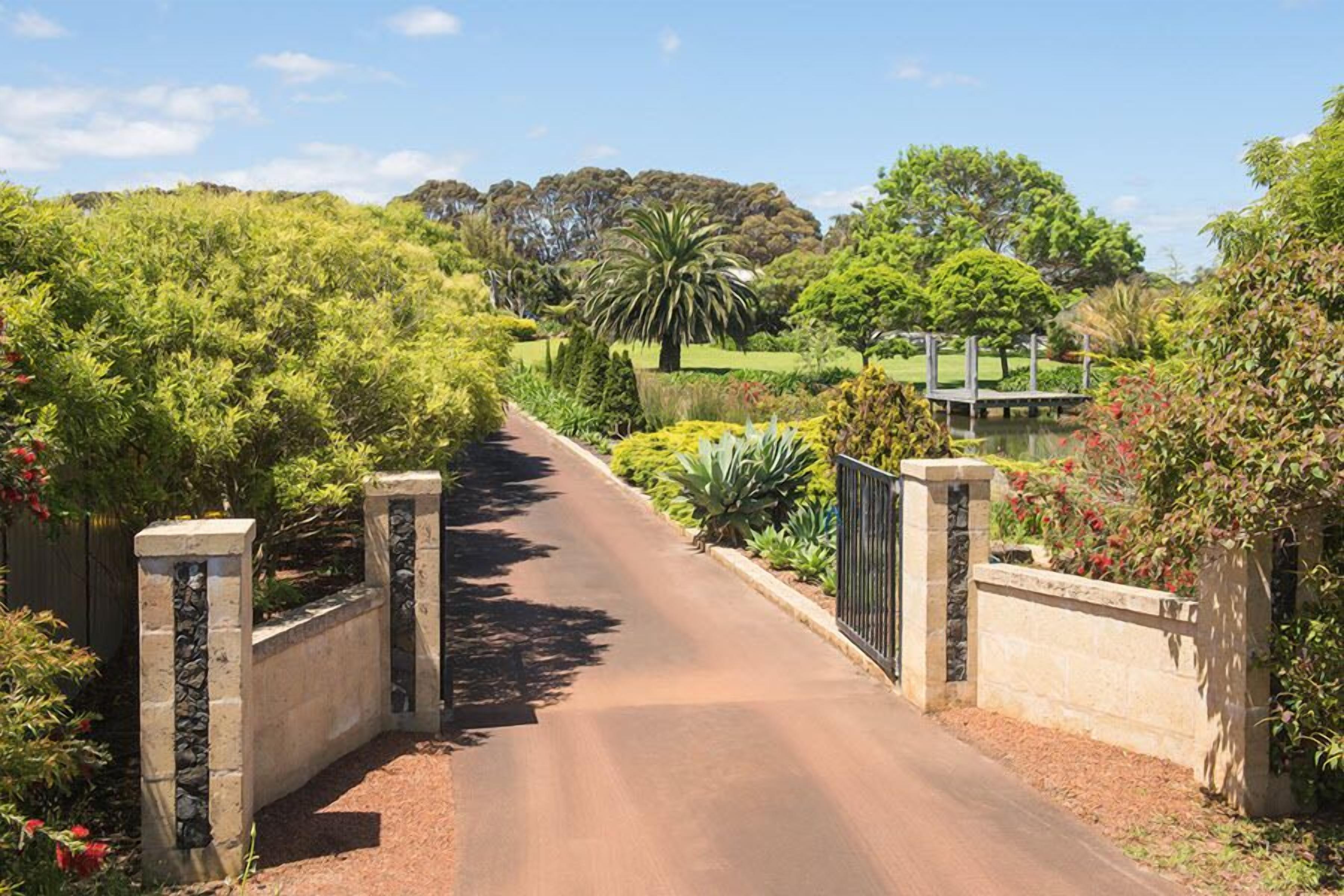 A paved pathway leading to a gated entrance surrounded by lush greenery and a small bridge over a water feature.