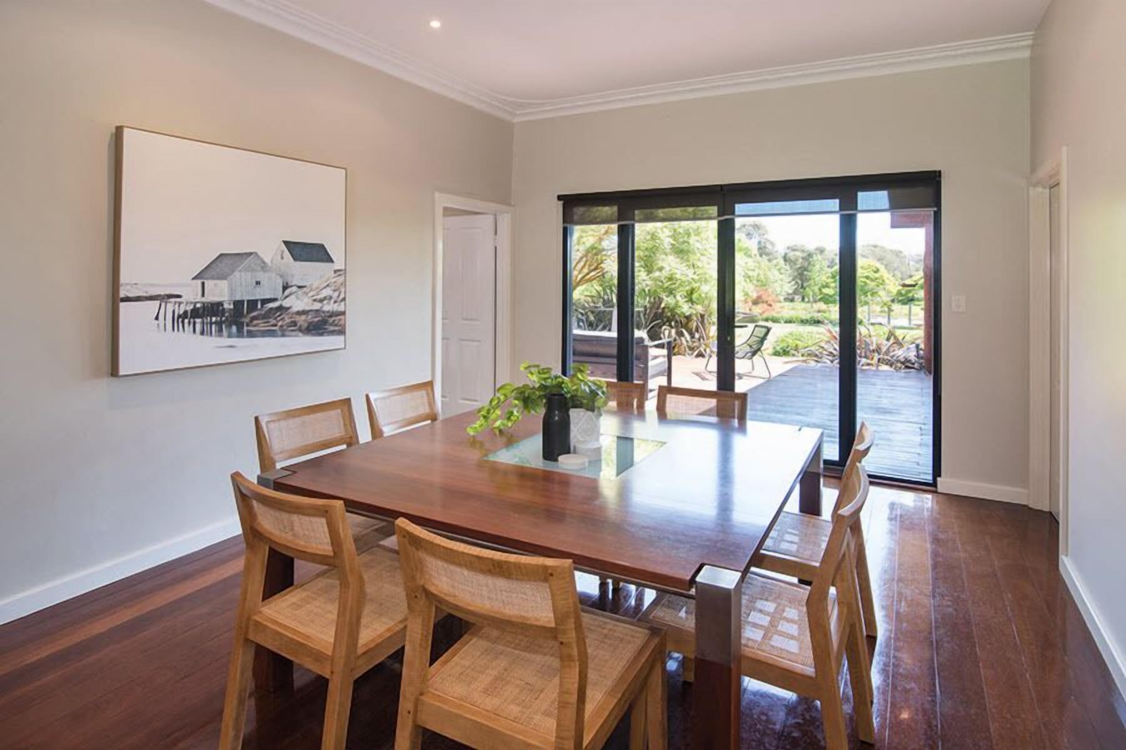 A dining area with a wooden table and chairs, a painting on the wall, and a view through a glass door to an outdoor area.