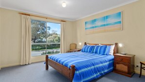 A bedroom with a large bed, a nightstand, a lamp, and a painting of a beach scene. - 22 Broadbeach Drive Carrickalinga (CARRICKALINGA)
