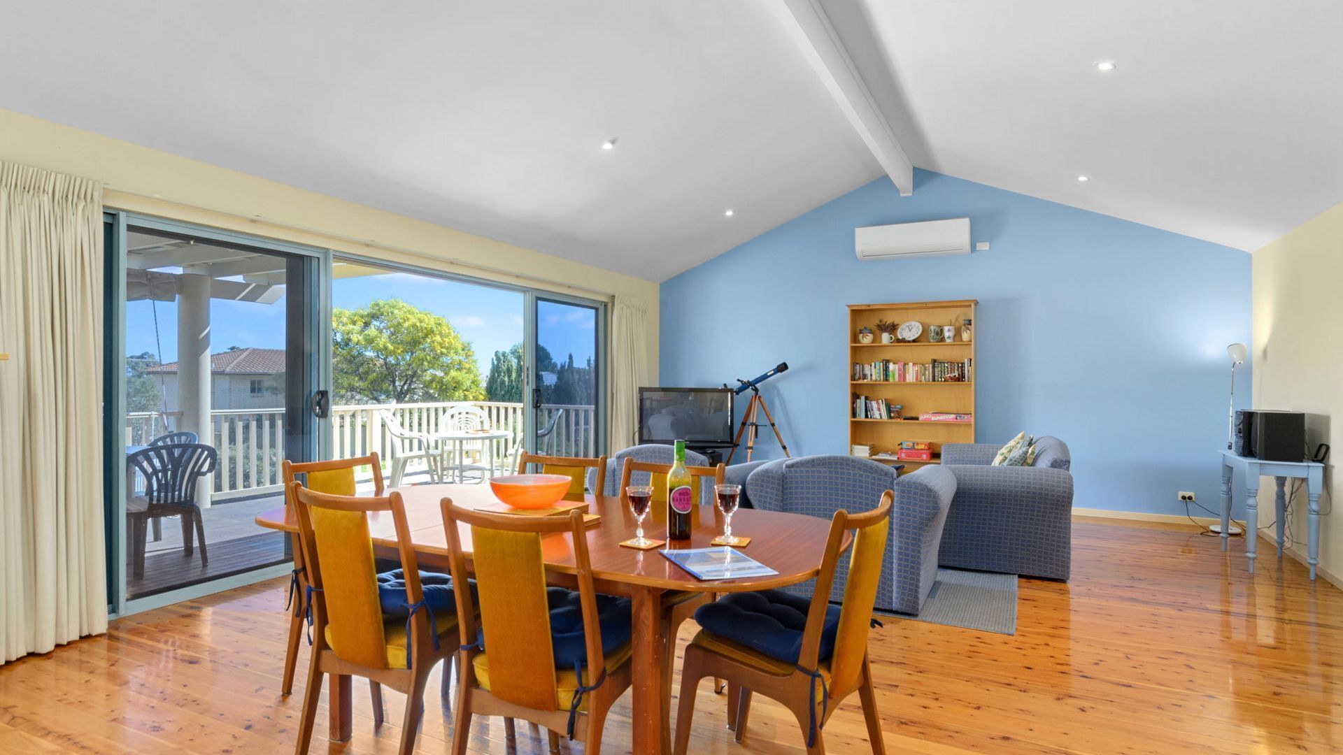 A spacious living area with a balcony, a dining table with chairs, a sofa, a bookshelf, and a telescope.