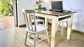 Una habitación con una mesa de madera, dos sillas blancas, un escritorio con una computadora y un cuadro enmarcado en la pared.