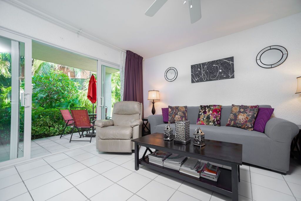 A modern living room with a grey sofa, a beige armchair, and a coffee table with magazines. There is a glass door leading to a balcony with a red towel hanging on it.