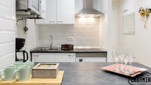 Une cuisine moderne avec des armoires blanches, un évier en acier inoxydable, une hotte intégrée et un plan de travail agrémenté d’une machine à café, de tasses et d’un panier de fruits.