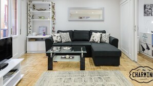 Un salon moderne comprenant un canapé d’angle, une table basse en verre, un meuble de rangement blanc pour appareils électroniques et une bibliothèque agrémentée de plantes décoratives.