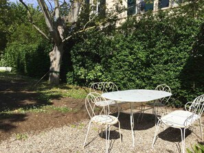 Une table ronde en métal, entourée de quatre chaises, posée sur du gravier et bordée de verdure, avec un arbre et des buissons touffus en arrière-plan.