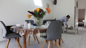 Un comedor con una mesa redonda cubierta con un mantel de plástico transparente, rodeada de sillas con patas de madera. Un jarrón con flores está sobre la mesa.