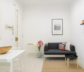 Un salon moderne avec une table blanche, un canapé noir agrémenté de coussins, un tableau encadré au mur et une petite étagère avec des plantes en pot.