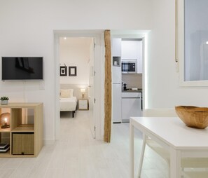 Une chambre d’hôtel moderne équipée d’une télévision, d’une table de chevet avec une lampe, d’un lit, d’une petite table de chevet avec une plante, d’une étagère en bois avec une lampe, et d’un coin cuisine avec un four à micro-ondes et un réfrigérateur.