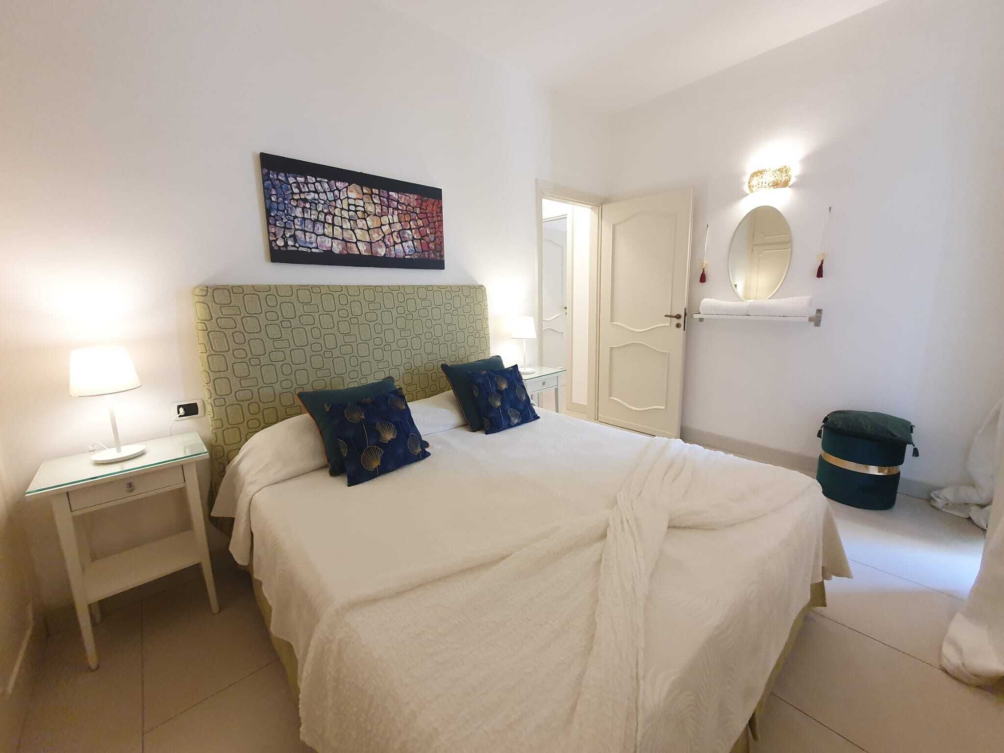 Habitación de hotel con cama, mesita de noche, lámpara, un cuadro, espejo y una puerta.