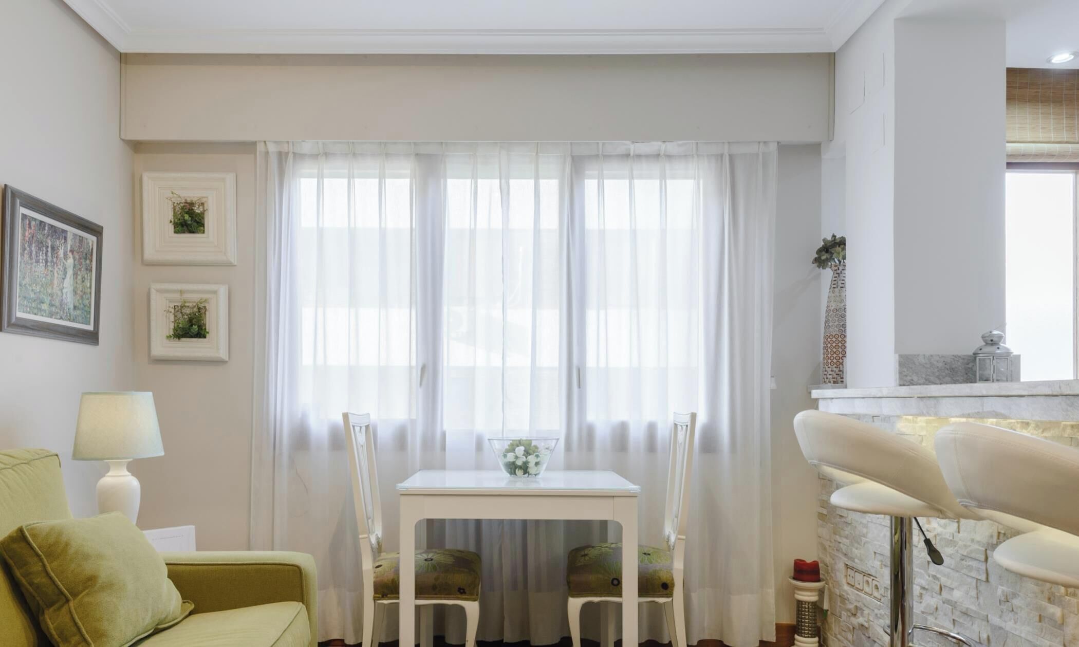 Een gezellige woonkamer met een groene bank, een witte tafel met stoelen, en een keuken met een achterwand van steen.
