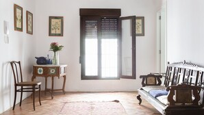 A room with a bed, a chair, a small table with a vase, and a window with blinds.