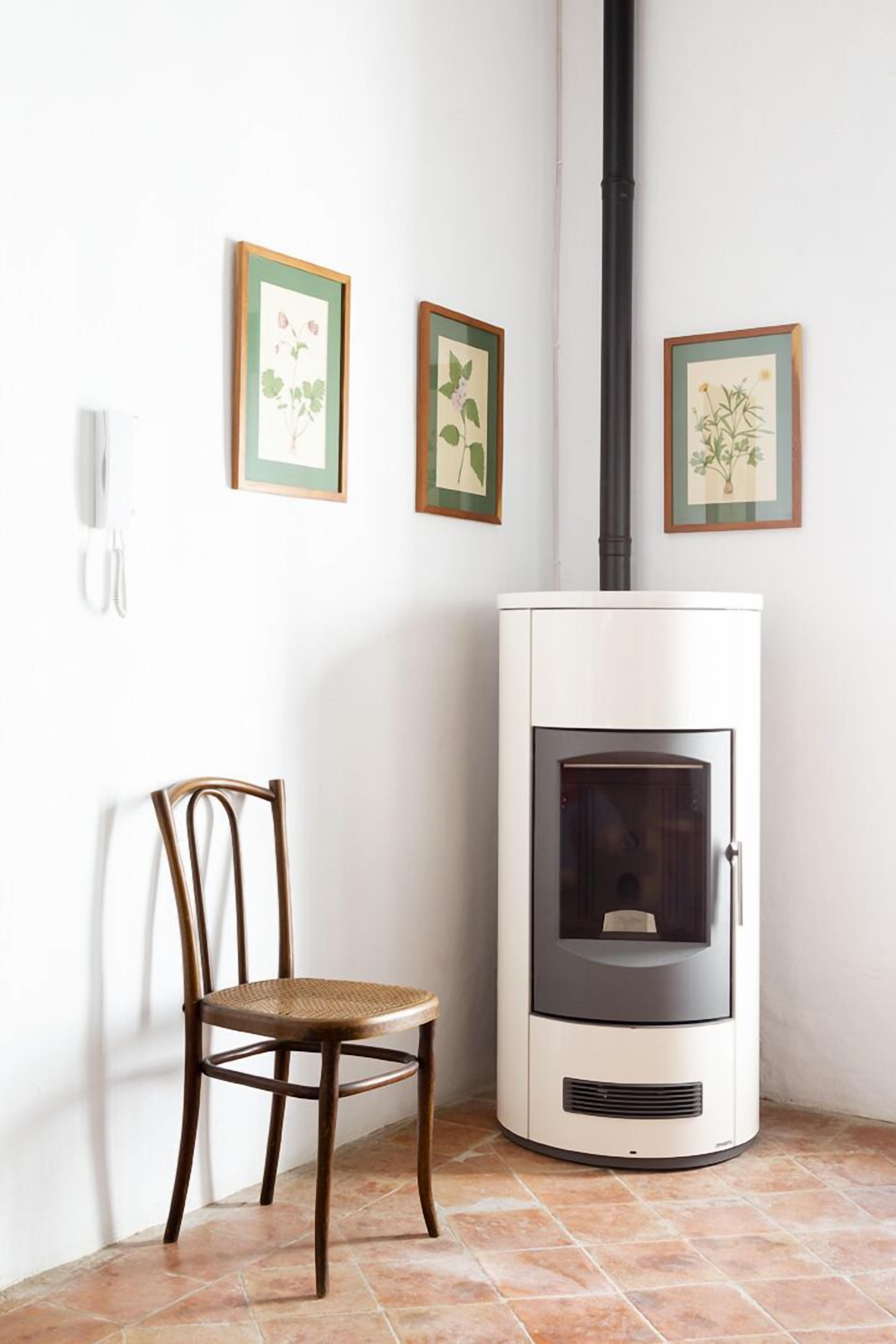 A corner room with a white wood-burning stove, a wicker chair, and framed botanical prints on the wall.