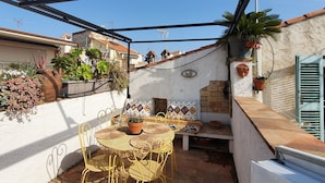 Un pati amb cadires de metall grogues i una taula rodona, envoltat de plantes en testos i una paret blanca amb un element decoratiu.