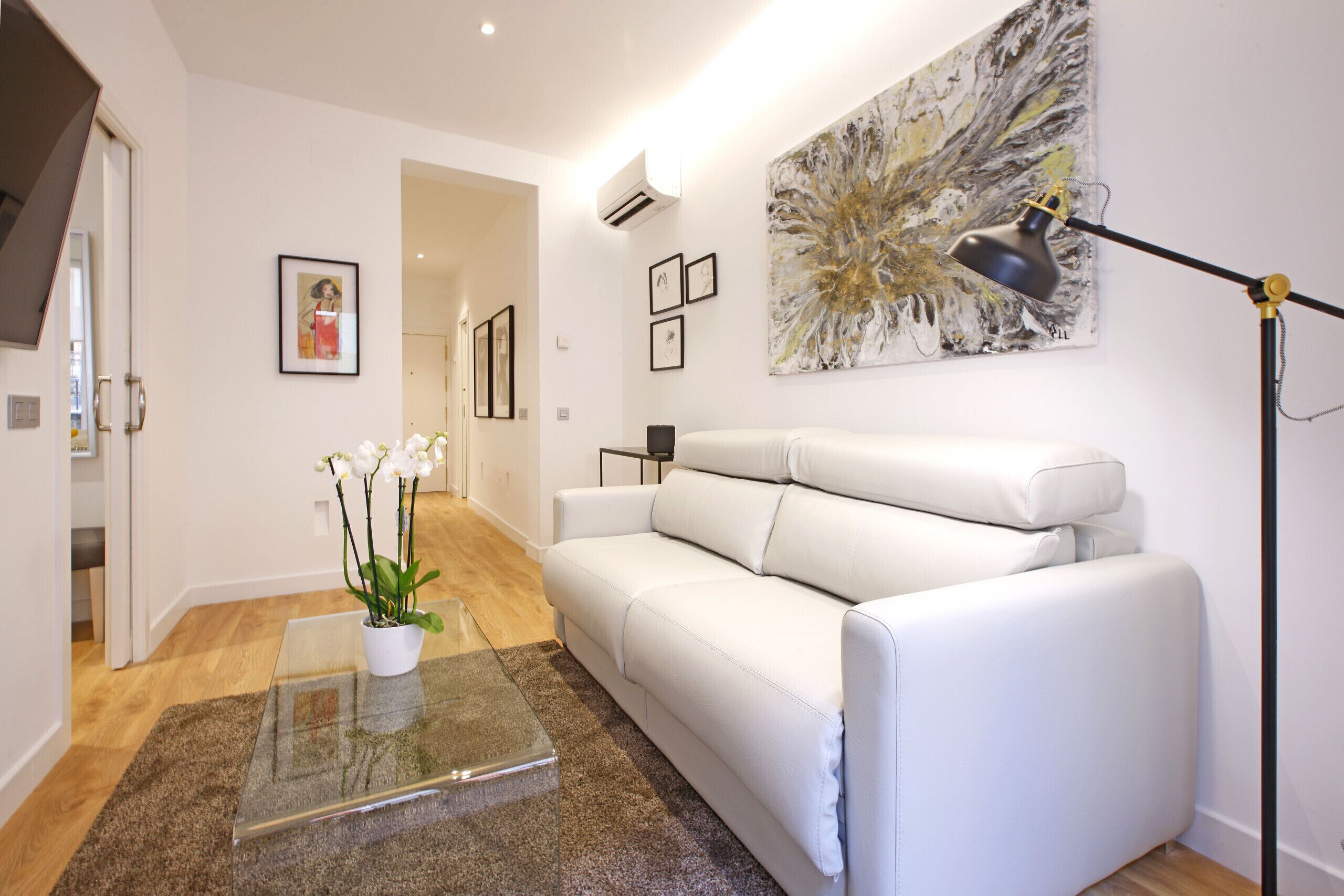 Ein modernes Wohnzimmer mit einem weißen Ledersofa, einem Glastisch mit Orchideen, einer Stehlampe und einem großen abstrakten Gemälde an der Wand.