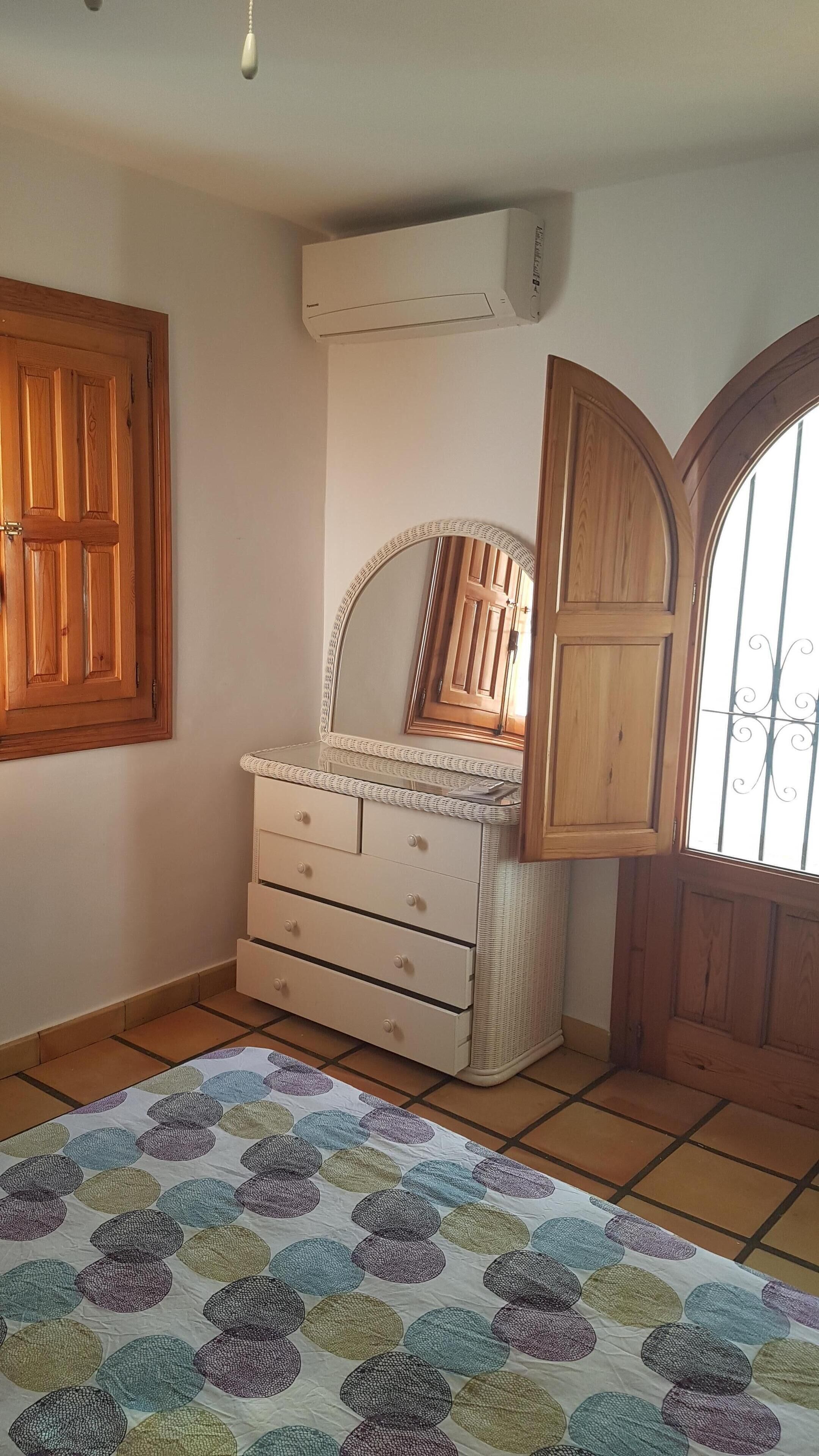 A bedroom with a wooden wardrobe, a dresser with drawers, a mirror, and a window with bars.