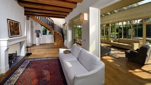 A spacious living area with a white sofa, a red patterned rug, a wooden staircase, and large windows allowing natural light.