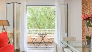A modern living room with a glass table, white chairs, a red sofa, and a balcony with a view of trees. - Madrid City - Two-bedroom apartment (Madrid)