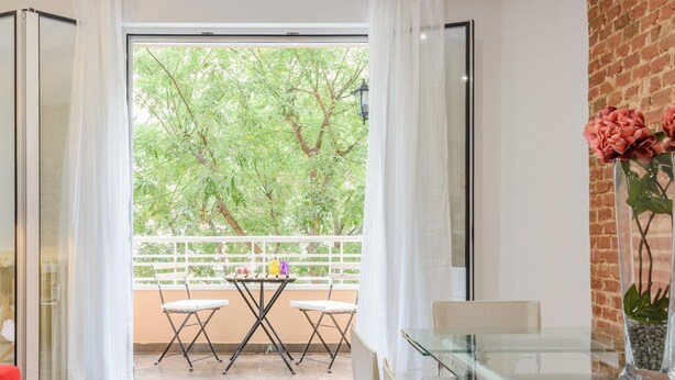 Uma sala de estar moderna com mesa de vidro, cadeiras brancas, sofá vermelho e varanda com vista para árvores.