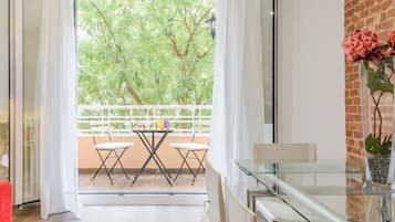 Een moderne woonkamer met een glazen tafel, witte stoelen, een rode bank en een balkon met uitzicht op bomen.