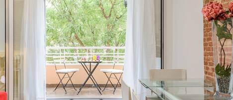 Un salon moderne avec une table en verre, des chaises blanches, un canapé rouge et un balcon donnant sur des arbres.