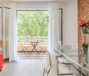 Un salon moderne avec une table en verre, des chaises blanches, un canapé rouge et un balcon donnant sur des arbres.