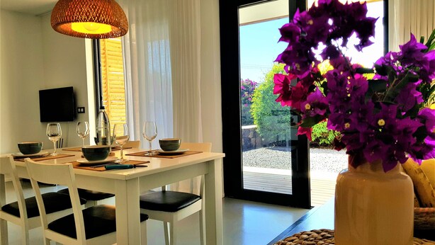 A dining area with a table set for two, a large vase with purple flowers, and a view of the outdoors through a sliding glass door. - Casa Cristina, new modern apartment in Lajares, Fuerteventura (Lajares)