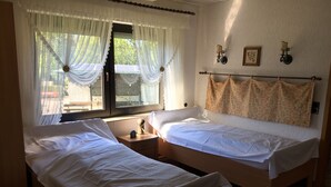 Une chambre avec deux lits simples, une petite table de chevet avec une lampe, une fenêtre avec des rideaux légers et un luminaire fixé au mur.