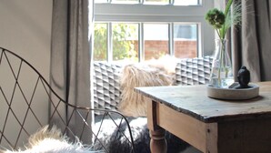 A room with a wooden table, a chair with a fur throw, a vase with a plant, and a window with a view of trees and a brick wall. - Stylish design apartment (Trier)