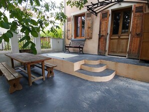 Un patio con una mesa y banco de madera, unos escalones que conducen a una puerta y una ventana con contraventanas.