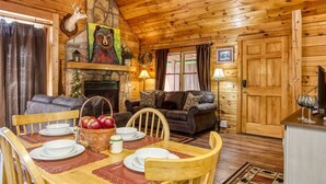A cozy cabin interior with a dining area, fireplace, and a living room with a couch and fireplace mantel.