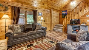 A cozy cabin interior with a wooden ceiling, a leather sofa, a small armchair, and a television on a wooden stand.
