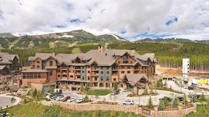 Un grand chalet à étages, dont la construction combine pierre et bois, se dressant sur un fond de montagnes et sous un ciel dégagé.