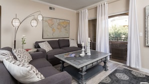 A living room with a dark wooden coffee table, a grey sofa with patterned cushions, a world map on the wall, and a view of the outside through a sliding door. - Workout room, yr round pool and spa, 2 Bd, 2 Bath, Gated, 9 AM-PM chk in-out (Phoenix)