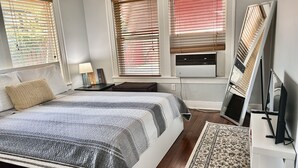 Ett sovrum med en säng, ett sängbord, en lampa, böcker, persienner, en luftkonditioneringsanläggning, en spegel och en matta.