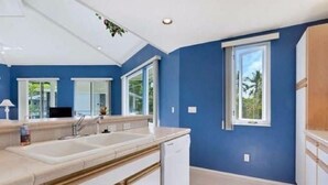 A modern kitchen with a large island, white cabinetry, and a view of greenery outside. -  Spacious, Luxurious, Tropical Townhouse Style Condo For A Truly Aloha Vacation (Kailua-Kona)