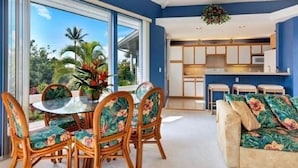 A modern living room with a glass dining table and chairs, a sofa with floral upholstery, and a view of a kitchen and outdoor greenery. -  Spacious, Luxurious, Tropical Townhouse Style Condo For A Truly Aloha Vacation (Kailua-Kona)