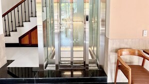 A modern elevator with glass doors, a staircase with wooden railings, and a chair in the foreground. - Soleil House Hostel (Hue)