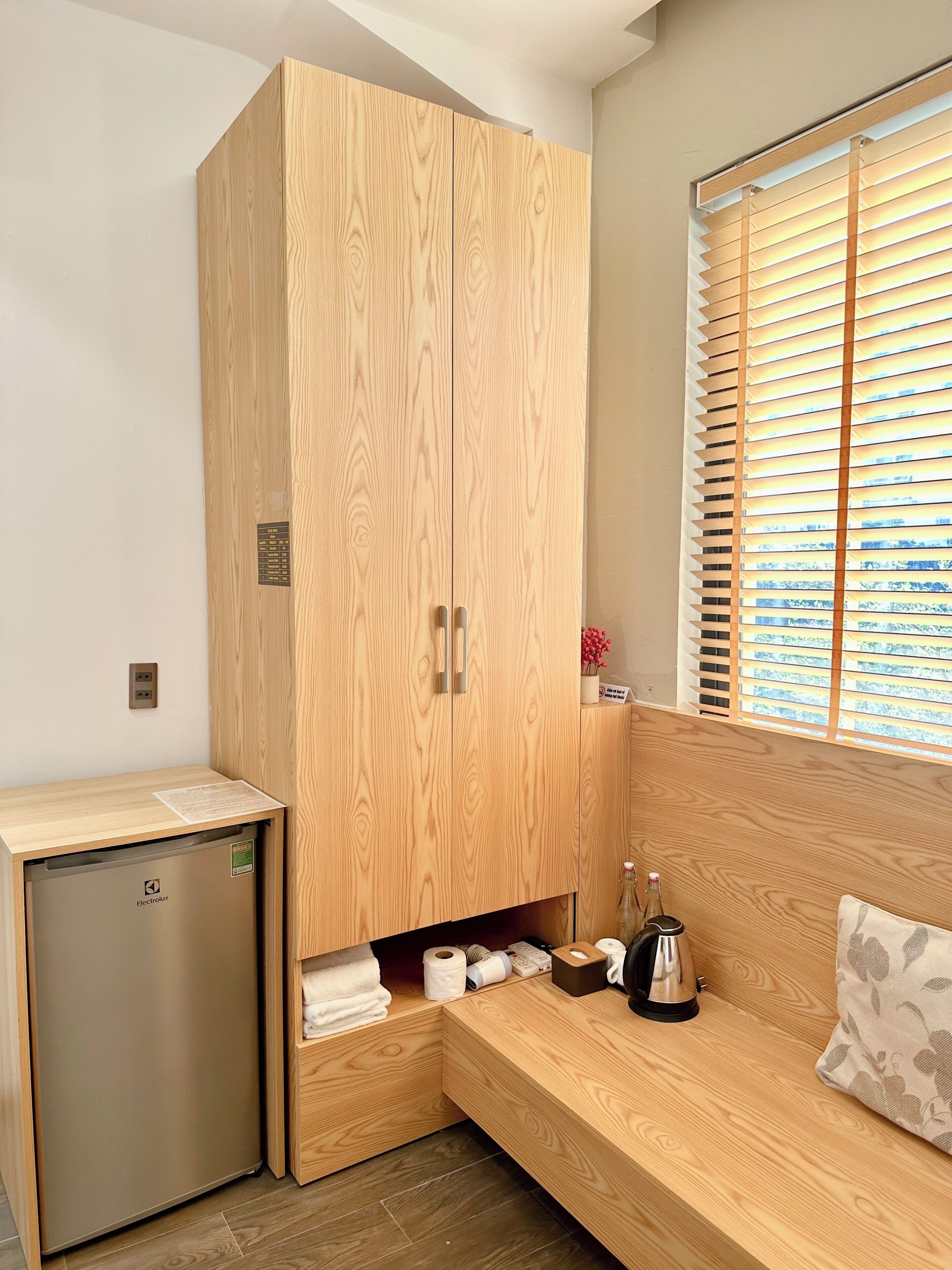 A compact room with a wooden wardrobe, a small refrigerator, a sofa, and a window with blinds.