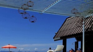 Zona de piscina con vista despejada al océano, una silla de descanso, una sombrilla roja y lámparas colgantes.