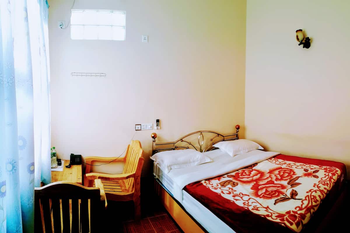 A single bed with a floral bedspread, a wooden chair, a small table with a bottle, and a window with a curtain.