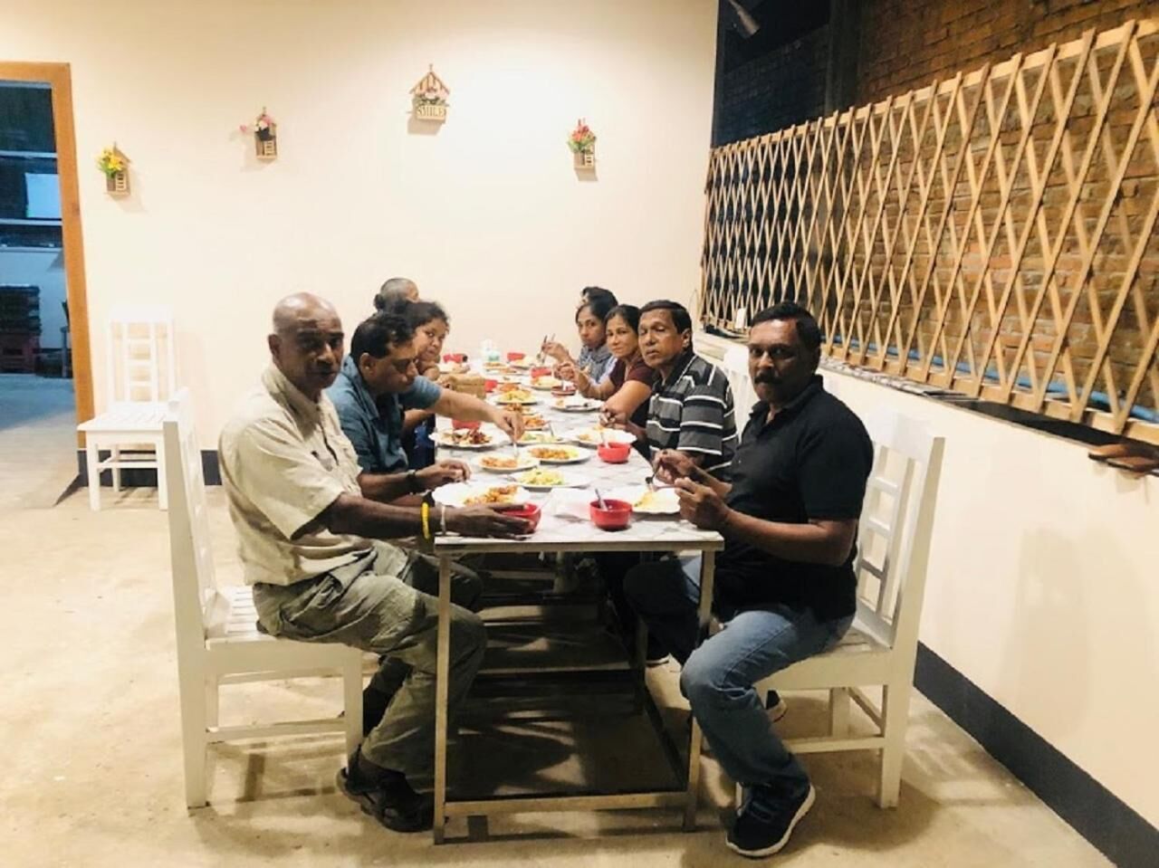 A group of people seated around a table in a room with a simple, functional design, featuring a plain wall and a lattice structure on one side.