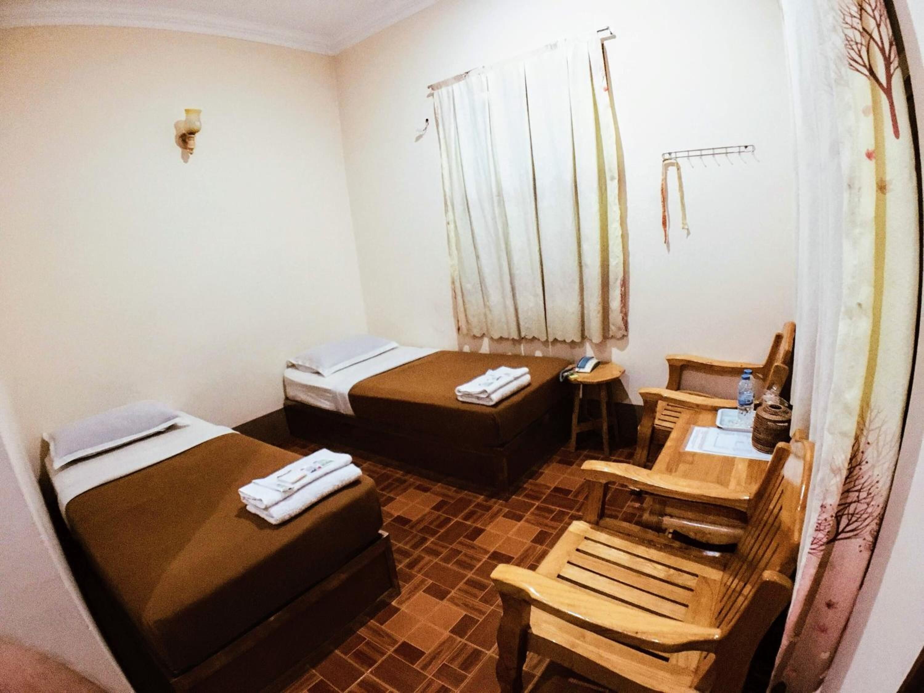 A hotel room with two beds, a small table with chairs, a window with curtains, and a wall-mounted light.