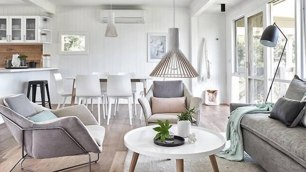 Ein modernes Wohnzimmer mit einem runden Couchtisch, einem grauen Sofa und einem grauen Sessel. Im Hintergrund ist ein Essbereich mit weißen Stühlen und einem weißen Tisch zu sehen.