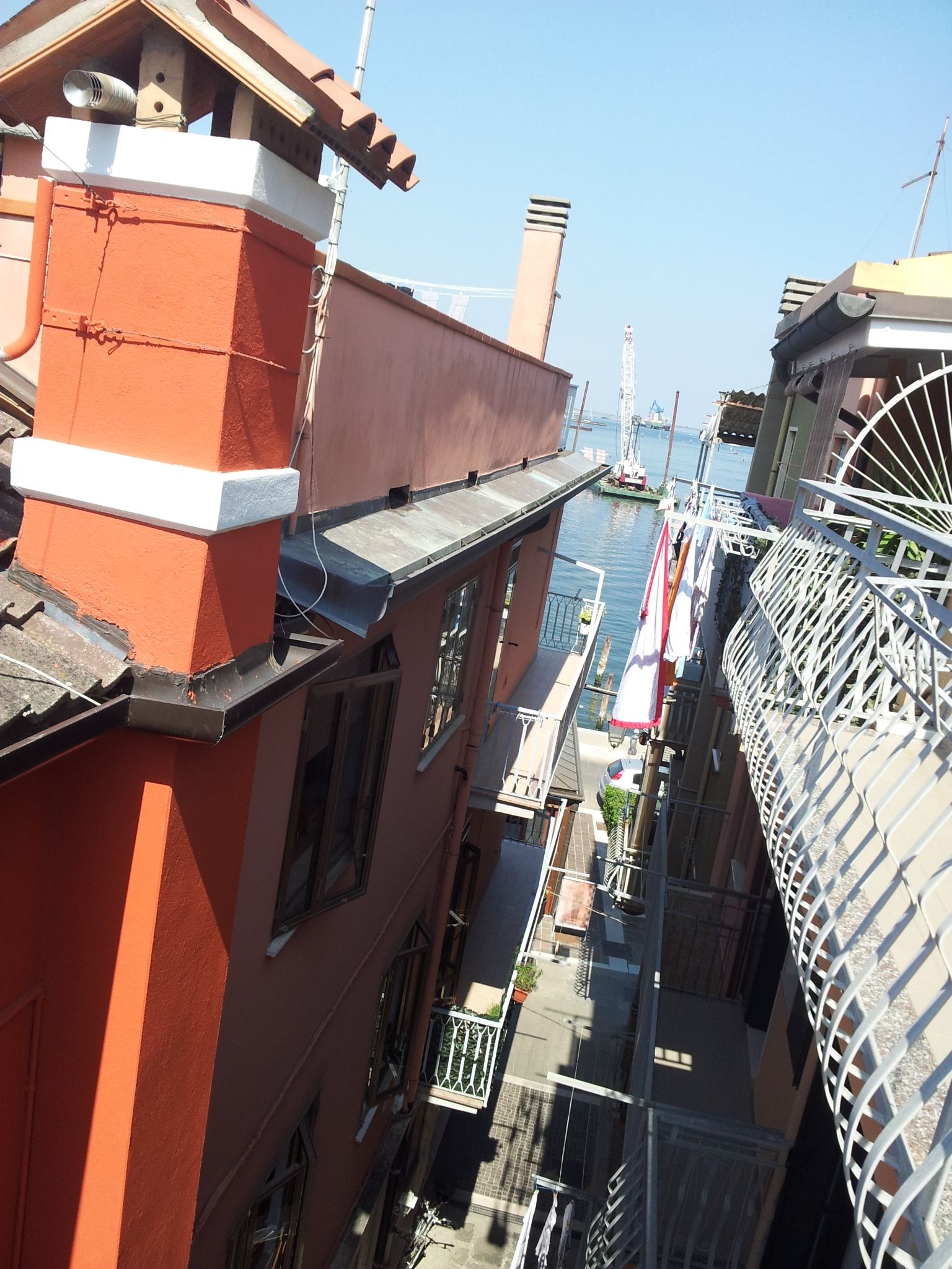 A narrow alleyway between buildings with a red brick facade, a white balcony railing, and a glimpse of a water body in the background.