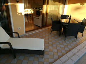 Un patio iluminado por el sol con una silla de descanso, una mesa de comedor con sillas y una cocina visible a través de una puerta abierta.