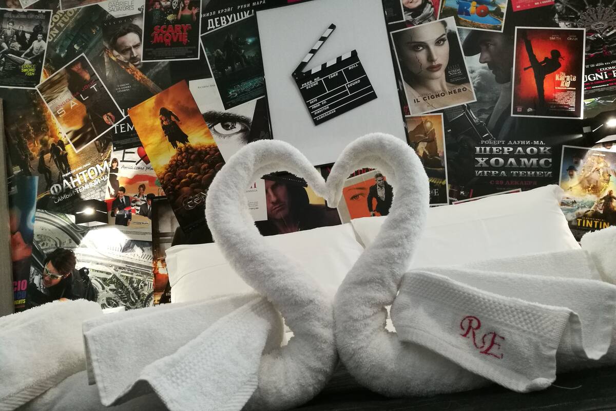 A room with a bed covered by a white blanket, a wall with movie posters, and a white towel with a heart-shaped design.